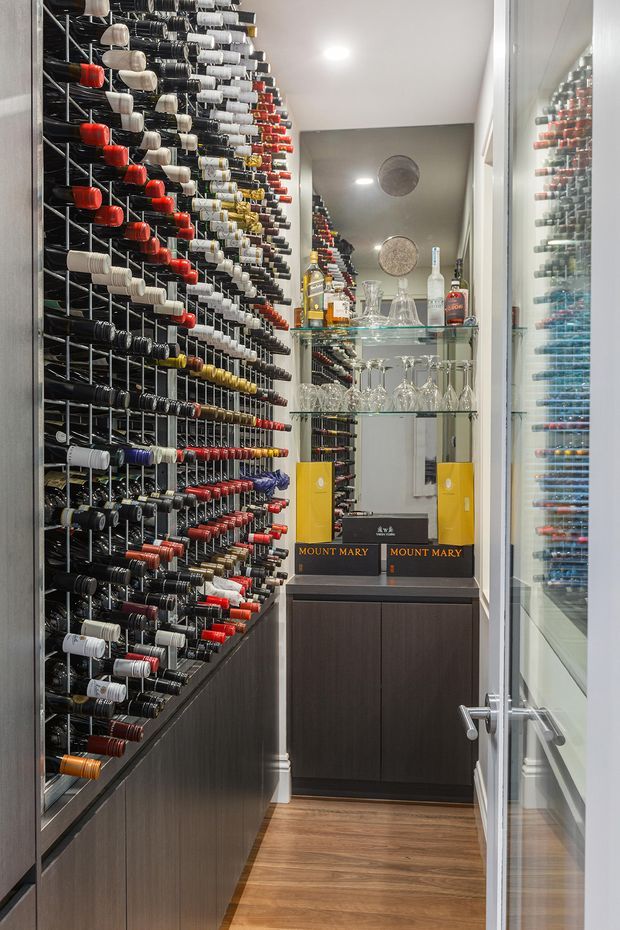 A narrow, compact wine storage room lined with extensive wall-mounted bottle racks. Dark wood cabinetry runs below the racks, while a mirrored and glass-fronted display cabinet holds wine bottles, decanters, and stemware. Warm timber flooring, recessed ceiling lighting, and a glazed door complete the refined wine cellar interior.