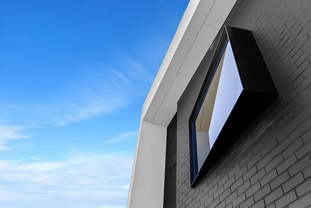 Low-angle exterior architectural detail showing a contemporary building facade. A dark grey brick wall is paired with crisp white geometric cladding and a projecting black-framed window or box feature. The image emphasizes angular composition, clean lines, and strong contrast against a blue sky.