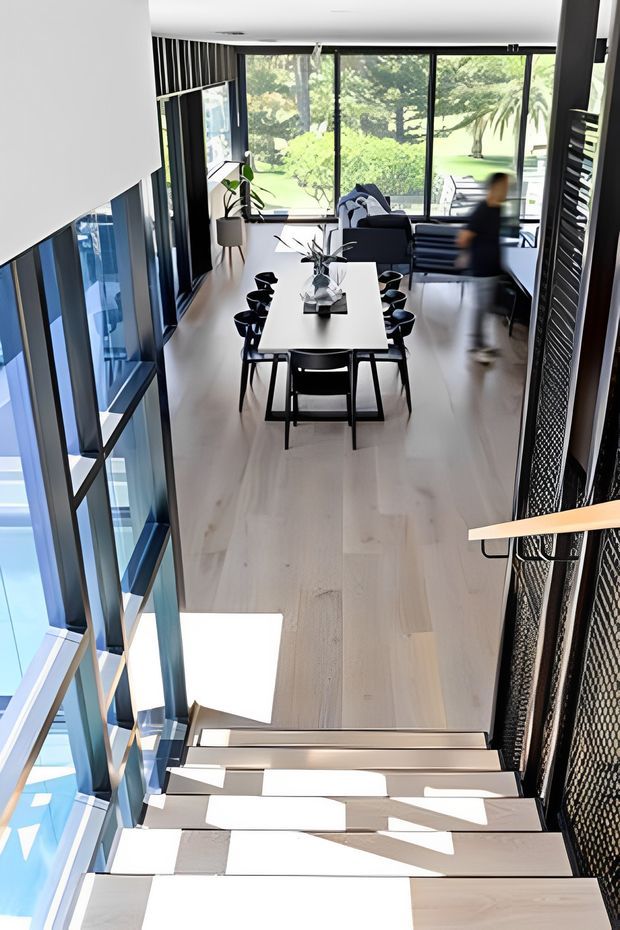 A bright, modern open-plan interior viewed from a stairway, looking down toward a dining area and living space. The room features a long light timber dining table with black chairs, extensive floor-to-ceiling glazing overlooking greenery, black-framed windows, pale timber flooring, and a sculptural black mesh stair balustrade. Strong sunlight creates geometric shadows across the stairs and floor.