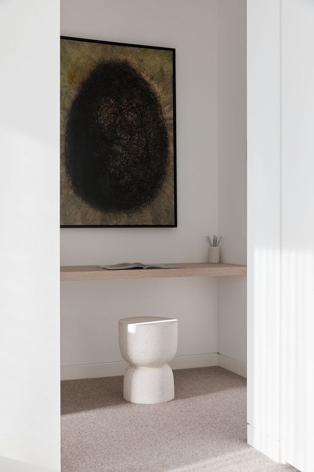 A minimalist interior nook with a pale painted wall, built-in light timber floating shelf, framed abstract artwork, and a small white sculptural stool or side table. The space has soft neutral carpeting, white architectural trim, and strong natural light filtering through vertical blinds.