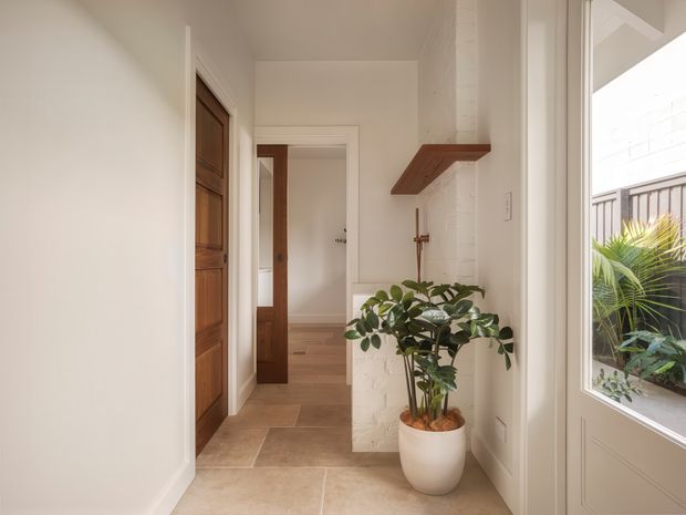 Bright, warm-toned residential hallway or entry passage with white painted walls, a tiled floor, timber panelled doors, and a white-painted brick fireplace or projecting wall feature with a simple timber mantel shelf. A potted indoor tree adds greenery, while glazed exterior doors or windows bring in daylight and show a balcony or garden area beyond.