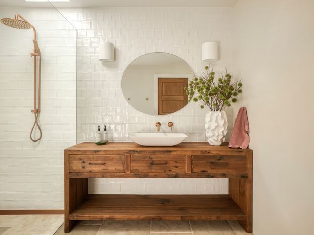 Bright, warmly styled bathroom featuring a walk-in shower with white glazed subway tile, a freestanding white vessel basin on a substantial rustic timber vanity, round wall mirror, brushed brass shower and tapware, and soft decorative accents. The timber vanity and natural accessories contrast with the light neutral finishes.