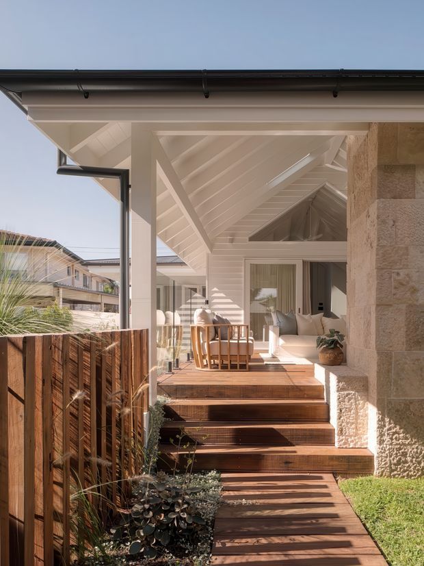 A bright architectural exterior view of a covered outdoor deck and entry terrace. Broad timber steps lead up to a partially enclosed veranda with timber decking, white-painted structural framing, exposed pitched roof rafters, large glazed openings, and a light natural-stone wall. The space contains a small dining setting and cushioned seating, with timber screening and landscaped planting alongside. Neighbouring residential buildings are visible beyond the boundary.