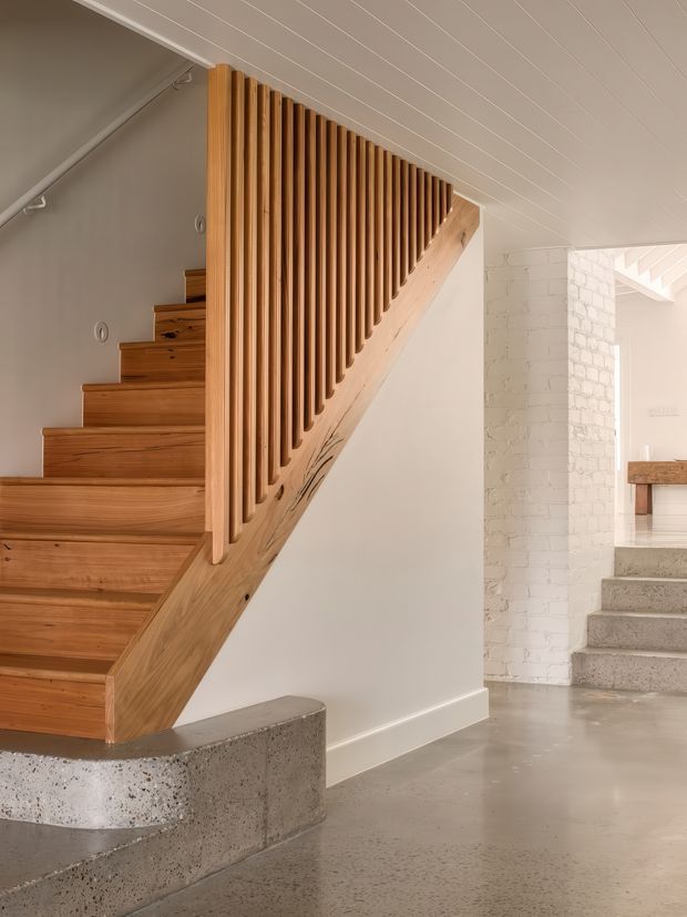 Interior view of a contemporary stairway with warm timber treads and a sculptural timber slatted balustrade. The stairs rise beside a polished concrete base and lead into a bright open-plan interior with white-painted brick walls, concrete flooring, and a glimpse of a kitchen or living area beyond.
