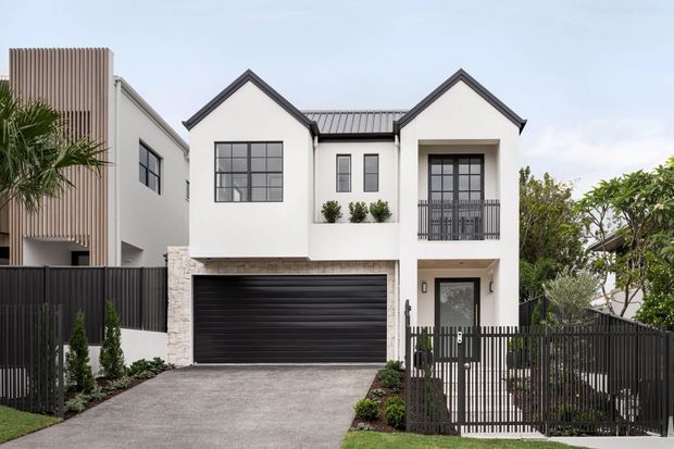 Front exterior view of a contemporary multi-storey townhouse with a symmetrical white rendered facade, two steep gable roof forms, dark metal roofing, black-framed windows and doors, a double garage, and a fenced driveway entry. Vertical timber-look screening is visible on the neighbouring or attached side facade, with restrained landscaping along the frontage.