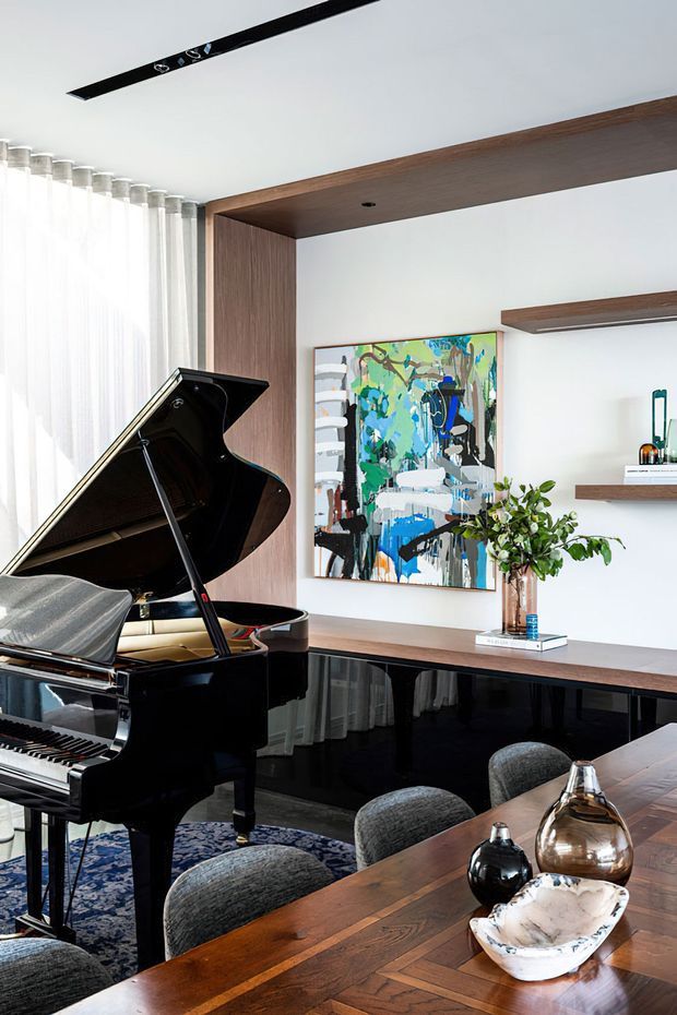 Refined open-plan living and dining space featuring a glossy black grand piano, a long timber dining table with upholstered chairs, dark wood built-in shelving and cabinetry, sheer full-height curtains, contemporary abstract artwork, and sculptural accessories. The room has a bright, polished, warm-modern aesthetic with strong timber and black accents.