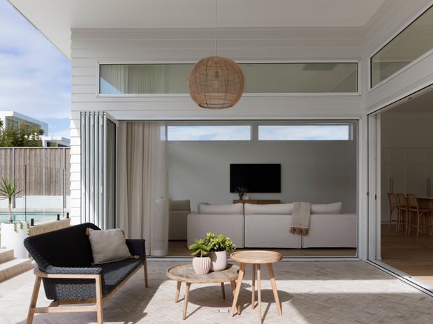 A bright, contemporary covered outdoor living area opens through large glazed folding doors into a minimalist living room. The space features white horizontal wall cladding, pale tiled flooring, woven black lounge seating, light timber coffee tables, a neutral sofa, a wall-mounted television, and a woven pendant light. A pool and neighbouring urban buildings are visible beyond the left side.