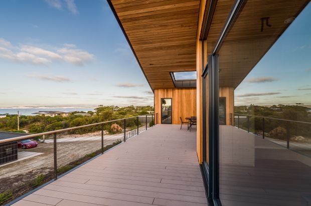 A contemporary elevated timber-clad house with a broad covered deck overlooking a coastal or bayside landscape. The deck features pale linear decking, slim dark metal-and-glass balustrades, warm timber soffit and wall cladding, large black-framed glazed openings, and a roof skylight. The reflective glazing mirrors the deck and surrounding view.