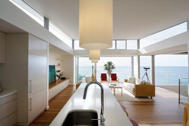 A bright, open-plan coastal interior combining a streamlined kitchen with a living area overlooking the ocean. The kitchen features tall pale cabinetry, a long white island with an integrated stainless-steel sink and curved tap, and warm timber flooring. Floor-to-ceiling glazing and clerestory windows create strong indoor-outdoor connection, while the living zone contains a light sofa, timber-framed seating, a coffee table, and large cylindrical pendant lights. A balcony with glass balustrading is visible beyond the living area.
