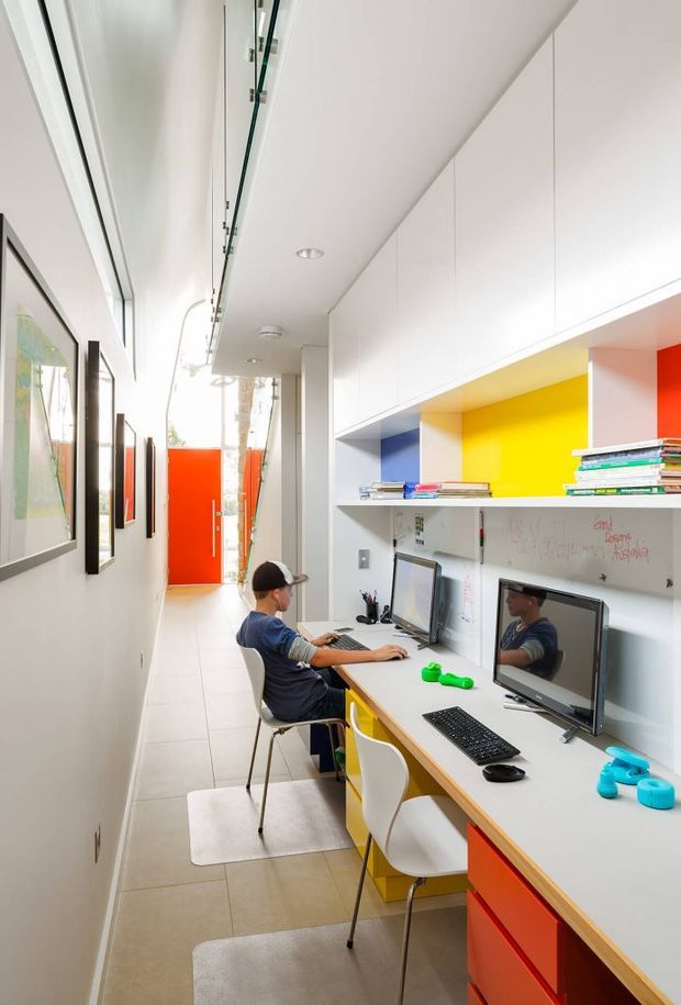 A bright, narrow contemporary home office or study corridor with a long built-in workstation. White overhead cabinetry, open shelving, colorful recessed niches in blue, yellow, and red, multiple desktop computers, and white chairs line one side. Framed artwork hangs along the opposite wall, while a vivid red door or panel anchors the far end. The space has a clean, playful, highly functional feel with tiled flooring and bright natural light.
