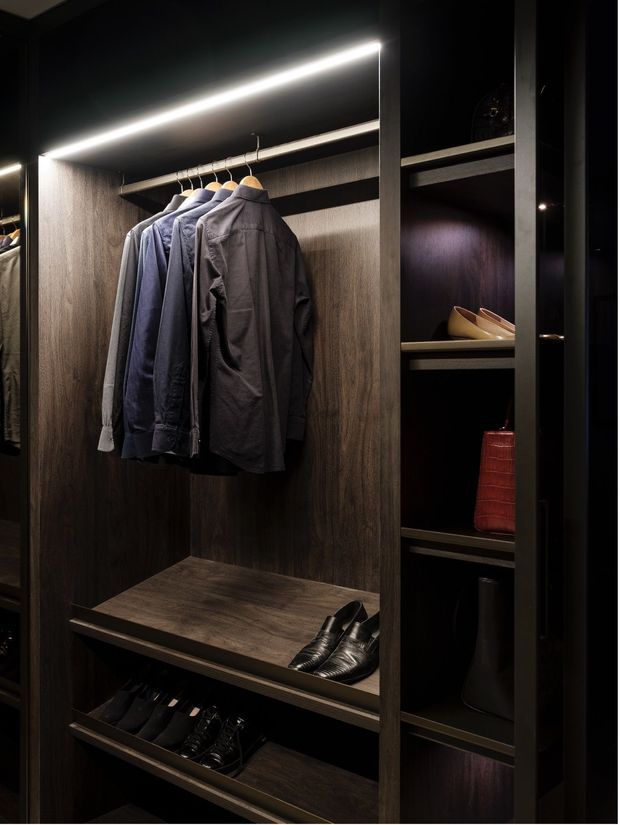 A dark, refined walk-in wardrobe with deep wood-grain cabinetry, open hanging rails, integrated linear LED lighting, shelves, shoe storage, folded accessories, and hanging jackets. The design has a moody, luxurious and highly organised appearance.
