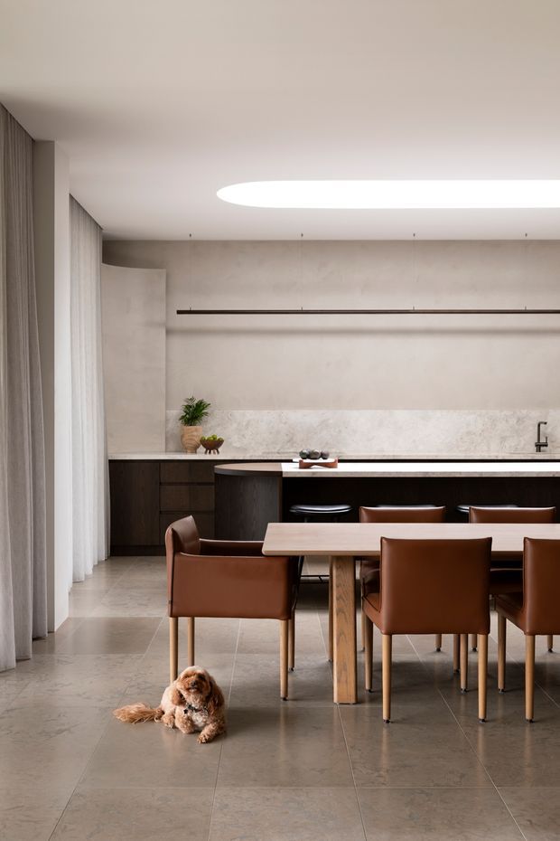 A refined open-plan dining and kitchen interior with a long timber dining table, brown leather dining chairs, dark built-in kitchen cabinetry, and a pale stone benchtop and splashback. Full-height sheer curtains line the left side, while a large recessed oval ceiling light creates a soft, contemporary focal point. The room features large-format stone-look floor tiles, warm neutral finishes, and minimal linear detailing.