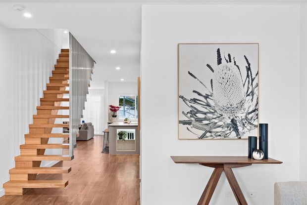 Bright contemporary open-plan interior featuring a floating timber staircase with slim vertical metal balustrades, pale timber flooring, white walls, and a narrow timber console table with angled supports beneath framed botanical artwork. A living area and kitchen zone are visible beyond the stair.