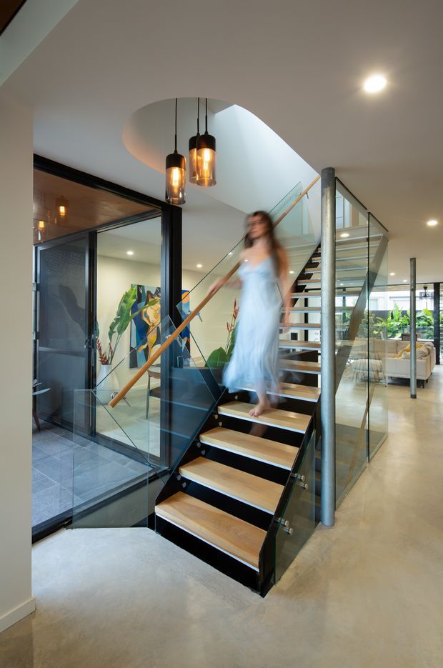 A contemporary interior focused on a sculptural open-riser staircase with warm timber treads, black metal stringers, glass balustrades, and a timber handrail. The stair rises through a curved ceiling opening beside a glass-enclosed room, with three suspended pendant lights overhead. Polished concrete flooring, exposed dark structural elements, indoor planting, and a bright living area are visible beyond.