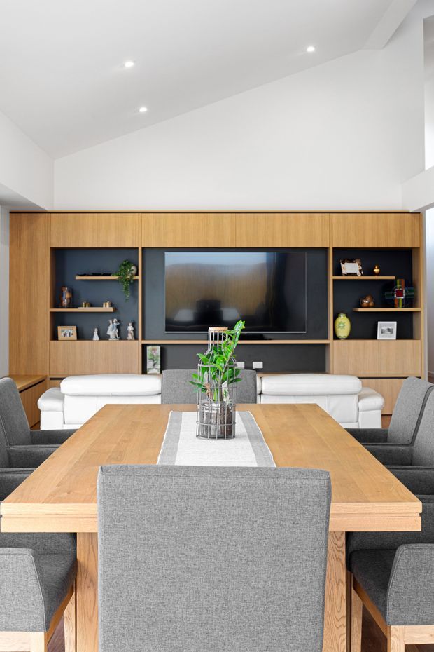 Bright contemporary open-plan dining and living area with a large light-wood dining table and grey upholstered dining chairs in the foreground. A white sofa sits behind the table, facing a wall-length built-in timber entertainment unit with dark shelving, a central television, and decorative objects. Recessed ceiling lights and a high white ceiling create an airy, clean interior.