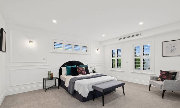 Bright, spacious bedroom with white decorative wall panelling, plantation shutters, recessed ceiling lights, wall sconces, neutral carpet, and a large upholstered bed with teal and patterned accent cushions. A bench and armchair provide additional seating.