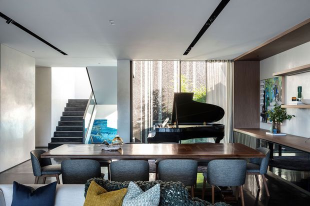 Open-plan contemporary interior combining a dining area and living space, with a dark wood dining table, upholstered dining chairs, a grand piano, built-in shelving, and a stairway leading to an upper level. Floor-to-ceiling glazing with sheer curtains brings in natural light, while recessed linear ceiling lighting and dark timber joinery add contrast.