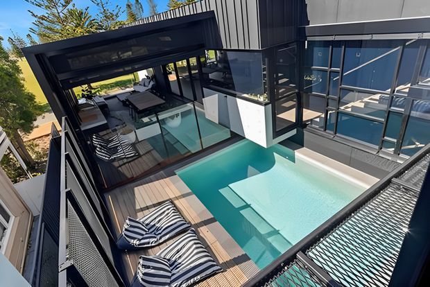 Elevated view of a contemporary multi-level home with a private turquoise swimming pool, timber decking, striped loungers, glass balustrades, dark metal framing, and a covered outdoor entertaining area. The pool terrace is integrated into the upper-level architecture and surrounded by urban residential landscaping.