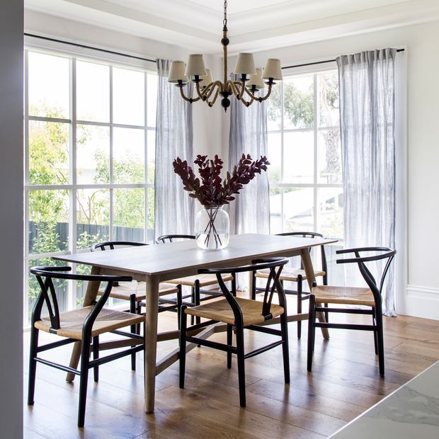 Bright, refined dining room with a large timber dining table, black-framed woven-seat chairs, a traditional chandelier, sheer curtains, and extensive multi-pane glazing. White walls with decorative moulding and warm timber flooring create an airy, classic interior.