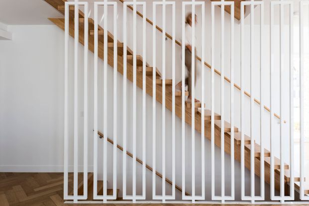 Bright contemporary stairway with a timber staircase and warm wood handrails set against white walls. A striking white vertical slatted balustrade or screen dominates the foreground, while herringbone timber flooring continues below. The image includes a partially obscured person on the stairs and strong natural light from glazing at right.