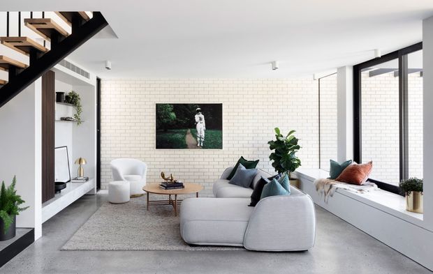 Bright contemporary living room with a large pale sectional sofa, armchair, round timber coffee table, and built-in window seat. The room features white-painted brick walls, polished concrete flooring, extensive black-framed glazing, minimalist track lighting, indoor plants, and a striking open-riser black timber staircase.