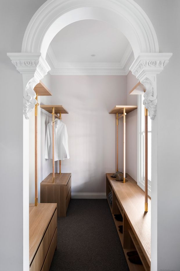 A narrow, elegant walk-in wardrobe framed by an ornate white arched opening with classical decorative mouldings and corbels. Warm timber open shelving, hanging rails, drawers and built-in benches line both sides, with clothing and shoes neatly arranged. A dark carpeted floor contrasts with the pale walls and white architectural detailing, while a tall window provides natural light.