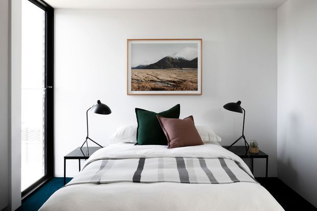 Bright, compact contemporary bedroom with a neatly made bed, white bedding and a striped throw, dark green and muted rose accent pillows, black bedside tables, and matching black angled desk lamps. White walls, dark flooring, and a tall black-framed glazed door or window create a clean, high-contrast interior. A framed mountain landscape artwork hangs above the bed.