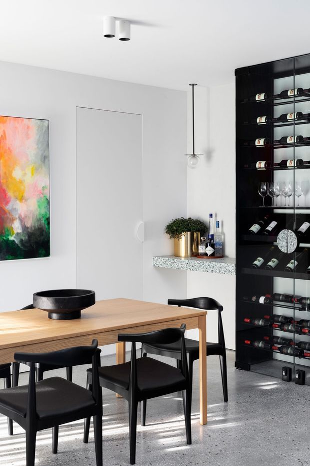 A refined contemporary dining area with a light timber dining table, black upholstered dining chairs, and a large black built-in wine storage cabinet displaying bottles and glassware. The room has white walls and ceiling, a polished terrazzo-like grey floor, minimalist lighting, a colourful abstract artwork, and a small stone-speckled serving counter with plants and drinks.