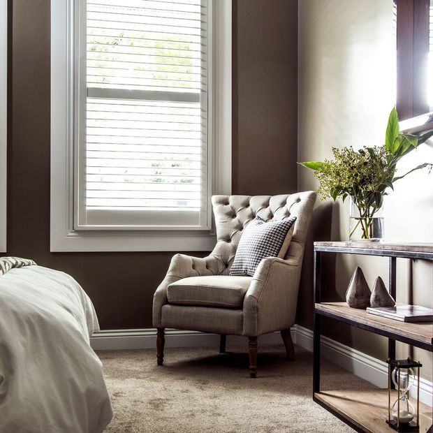 A warmly lit bedroom corner featuring a tufted upholstered armchair beside a tall window with white plantation shutters. A partially visible bed sits in the foreground, while a slim dark-framed console displays decorative objects and a vase of greenery. The room has dark taupe walls, light carpeting, and a refined, cozy interior.