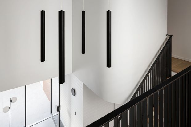 A minimalist contemporary stairwell viewed from an upper landing, featuring black metal balustrades, pale walls, timber flooring, and a cluster of long cylindrical black pendant lights suspended over the stair void. Glazing and a lower-level area are visible below.
