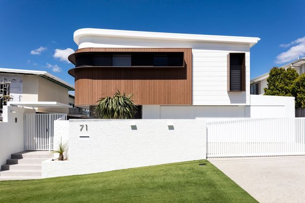 Contemporary two-storey residential facade with a curved white roof edge, crisp white horizontal cladding, warm vertical timber-look wall cladding, dark recessed glazing, and a white textured boundary wall with integrated house number. A manicured lawn, front steps, driveway, and simple spiky landscaping complete the suburban street frontage.