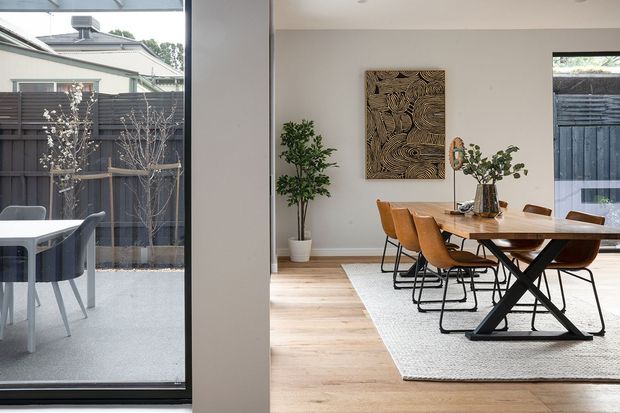 Bright contemporary dining area viewed through a large glazed opening, with a timber dining table, tan upholstered chairs, a neutral rug, pale timber flooring, indoor plants, and a carved wood wall artwork. A small adjacent outdoor or enclosed dining area is visible through the glazing on the left.