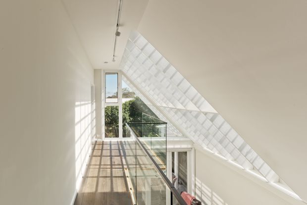 Bright contemporary interior landing or stair hall beneath a steeply sloping glazed roof. The space features white walls and ceiling, extensive translucent or glazed roof panels, a tall vertical window, and a glass balustrade overlooking the level below. Strong sunlight casts geometric shadows across the timber floor and walls, creating an airy, light-filled architectural atmosphere.