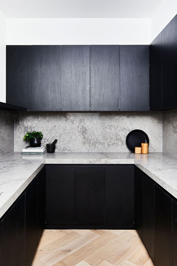 A compact, sophisticated galley-style kitchen with dark vertically grained cabinetry, pale grey veined stone benchtops and matching splashback, and light timber parquet flooring. The U-shaped layout includes extensive overhead and base storage, with minimal black and natural timber styling accents.