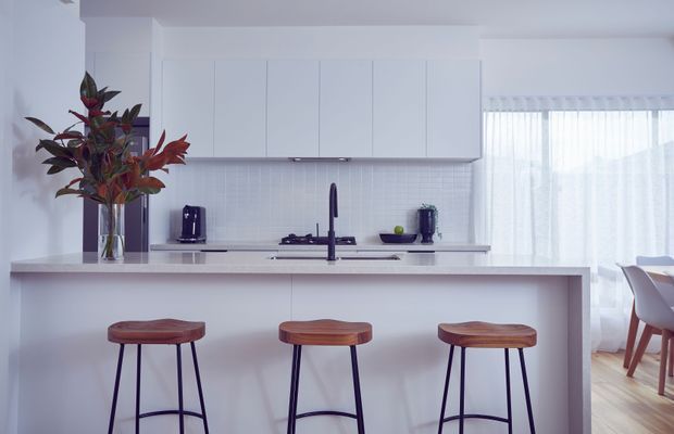 Bright contemporary white kitchen with a large island and three timber-topped bar stools. The space features white flat-panel cabinetry, a white tiled splashback, pale benchtops, dark tapware and appliances, timber flooring, and sheer curtains opening toward a dining area. A floral arrangement adds a small natural accent.