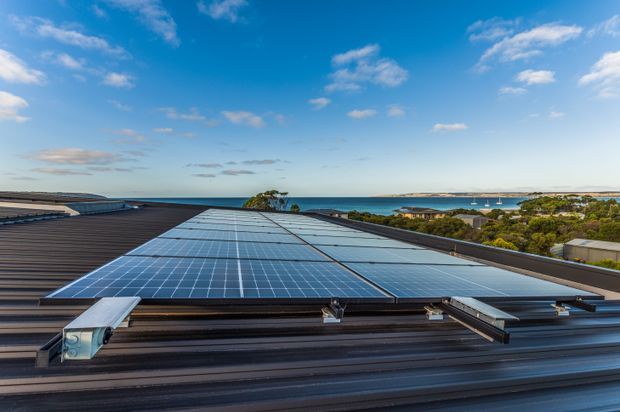 Aerial view looking across a large corrugated metal roof fitted with rows of photovoltaic solar panels and mounting rails. Beyond the roofline is a coastal or waterfront landscape with low-rise buildings, trees, calm blue water and a distant shoreline under a bright blue sky.