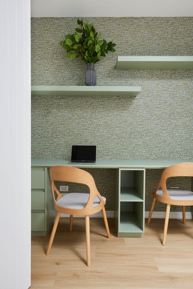 A compact home office viewed through a doorway, featuring a long pale sage-green built-in desk with open shelving and drawers, two light timber chairs with upholstered seats, and a laptop. The room has warm timber flooring, textured grey-green wallpaper, minimalist floating shelves, and a decorative vase with foliage. The palette is calm and muted with contemporary, Scandinavian-inspired styling.