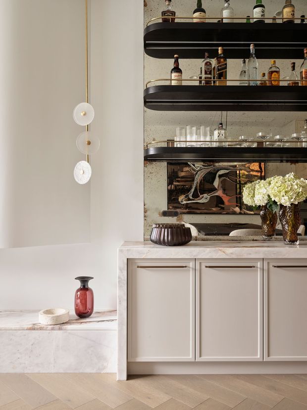 A refined home bar or drinks cabinet built into a pale, elegant interior. The lower section features cream shaker-style cabinetry with slim metal pulls and a pale marble benchtop, while the upper section has multiple dark floating shelves with brass rails, mirrored or reflective backing, liquor bottles, glassware, and decorative objects. A sculptural wall light, artwork, vase, flowers, and herringbone timber flooring add warmth and sophistication.