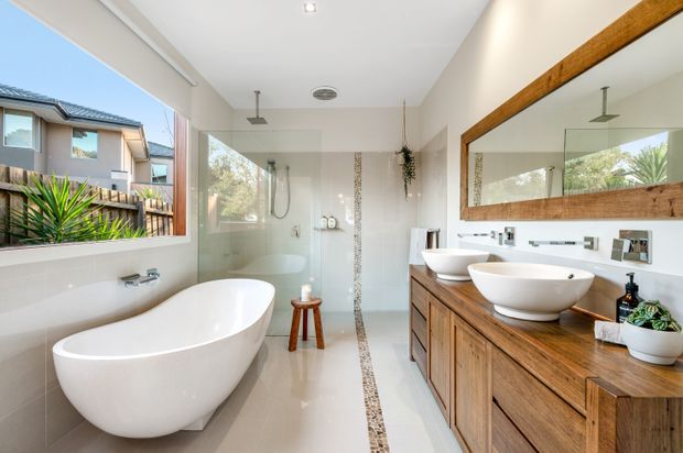 Bright contemporary bathroom with a freestanding white bathtub, frameless glass walk-in shower, double vessel basins, and a wide timber vanity. Large windows bring in natural light and overlook neighbouring residential landscaping. A full-width mirror with a substantial timber frame and a narrow decorative floor inlay add warmth and detail.