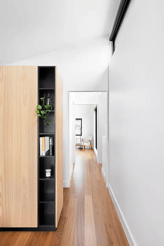 Bright minimalist hallway with white walls and ceiling, warm timber flooring, and a built-in light-wood partition featuring open black shelving with books and a trailing plant. A doorway and glimpse of a chair are visible at the end of the corridor.