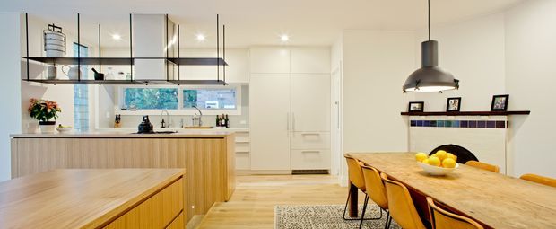 Open-plan kitchen and dining area with a warm contemporary fit-out. The kitchen features pale timber-look cabinetry, a light benchtop, integrated appliances, open shelving and a black-framed glass partition or display element overhead. A large timber dining table with mustard-yellow upholstered chairs occupies the foreground, while a simple linear shelf and artwork feature wall sit beneath a dark pendant light. Light timber flooring, white walls and abundant daylight create a bright, inviting space.