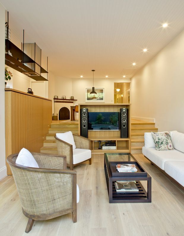 Warm, compact contemporary living room with a television and media console integrated into a timber built-in unit. The room includes a white sofa, woven upholstered armchairs, a dark rectangular coffee table, light timber flooring, timber cabinetry and stepped timber seating or platform elements. A raised dining or study area is visible at the rear, with open black-framed shelving overhead and recessed ceiling lighting. The interior has a clean, warm, mid-century-influenced character.