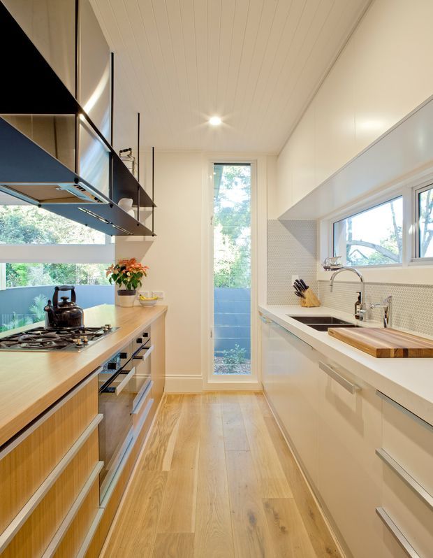Bright, narrow galley kitchen with white full-height cabinetry, pale timber flooring, a timber-topped cooking island, integrated appliances, gas cooktop and dark rangehood. The opposite wall has a white stone-look benchtop, undermount sink, chrome tapware, white small-format tiled splashback and a timber chopping board. Large windows and a glazed door provide abundant natural light and views to greenery. The ceiling is lined with white painted boards.