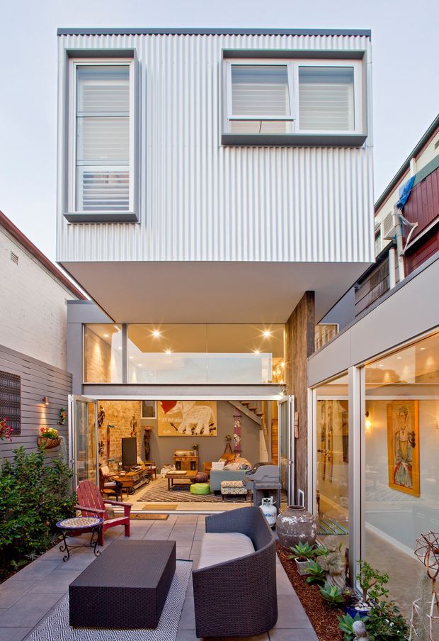 A narrow urban courtyard framed by a contemporary multi-level home. The image looks toward a glazed living area beneath a cantilevered upper storey clad in pale vertical corrugated metal, with clerestory-like windows and strong indoor-outdoor connection. The courtyard is furnished with woven outdoor lounge seating, a low coffee table, tiled paving, planter beds, and decorative lighting. Warm interior lighting, exposed brick, timber-toned finishes, and large glazed openings create an inviting contemporary atmosphere.