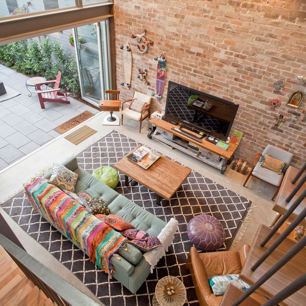An eclectic loft-style living room viewed from an upper level, with a large exposed brick feature wall, double-height glazing opening to a paved courtyard, timber-framed windows, and a mezzanine stair railing in the foreground. The room contains a pale green sofa with a colourful woven throw, leather armchair, patterned area rug, timber coffee table, media console and television, plus assorted decorative wall objects. The space is bright and characterful, combining industrial architectural elements with colourful vintage-inspired furnishings.