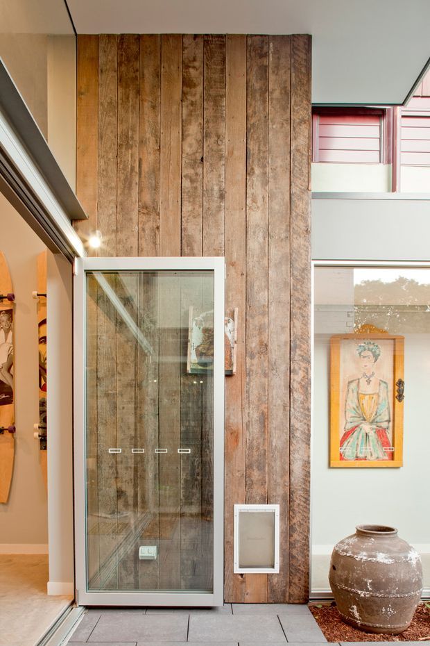 Interior view of a contemporary entry or hallway featuring a tall vertical reclaimed timber feature wall, a full-height glass door, pale walls, grey tiled flooring, and high clerestory-style glazing. A large decorative ceramic urn and framed artwork add warm accents. The space appears bright, curated, and materially expressive.