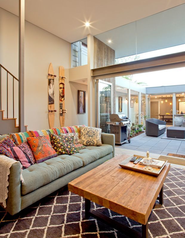 A bright, open-plan living room with a high ceiling and expansive glazing opening onto a covered courtyard or outdoor entertaining area. The room features a pale green upholstered sofa layered with colourful patterned cushions, a large rustic timber coffee table, a dark geometric rug, exposed stair detailing, decorative wall-mounted boards, and contemporary outdoor furniture beyond the glazing. Warm recessed and pendant lighting creates an inviting atmosphere.