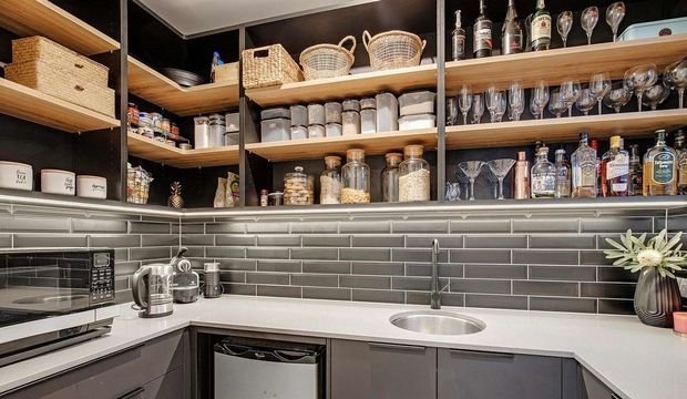Compact, well-organised butler's pantry or bar-preparation area with dark cabinetry, open timber shelving, woven storage baskets, glassware and bottles. A white benchtop incorporates a round stainless-steel sink, with a dark grey subway-tile splashback and integrated appliances.