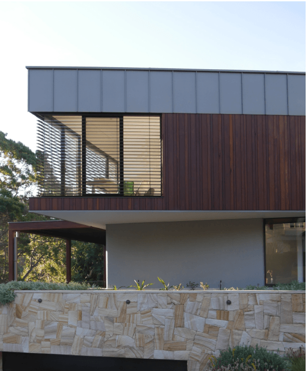 Exterior view of a modern two-storey house with a flat roof, dark vertical timber cladding, pale rendered walls, broad overhanging eaves, and a screened upper-level opening with horizontal louvres. A covered outdoor area and stone-clad retaining wall are visible in the foreground, with mature landscaping around the building.