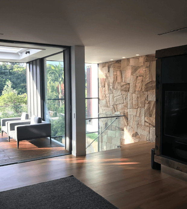 A contemporary interior showing a timber-floored living area beside a glazed sunroom or enclosed outdoor lounge. Full-height glazing opens to leafy greenery, while a rugged natural stone feature wall, glass balustrade, recessed lighting and dark built-in media joinery define the space. The room is bright and connected to the landscape, with a warm, natural material palette.